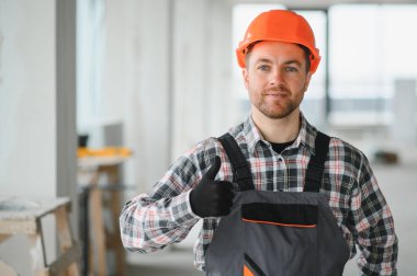Turuncu kasklı ve tulumlu kendine güvenen müteahhit bina içinde inşaat halindeyken iyi niyet göstergesi gösteriyor.