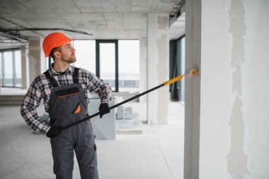 İnşaat işçisi koruyucu kask ve tulum giyiyor, uzun bir silindir kullanarak duvarı boyuyor, tadilat sırasında...