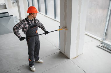 İnşaat işçisi koruyucu kask ve tulum giyiyor, uzun bir silindir kullanarak duvarı boyuyor, tadilat sırasında...