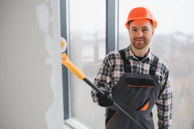 İnşaat işçisi koruyucu kask ve tulum giyiyor, uzun bir silindir kullanarak duvarı boyuyor, tadilat sırasında...
