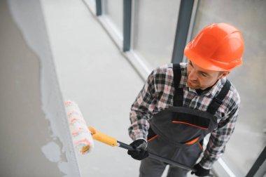 İnşaat işçisi koruyucu kask takıyor ve iş tulumları giyiyor. Duvara boya silindiri kullanarak astar takıyor ve son boyaya hazırlıyor.