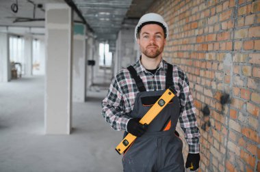 Miğfer takan inşaat işçisi, ruhsal bir seviyede duruyor. Tamamlanmamış bir binayı dikkatle ve titizlikle inceliyor.