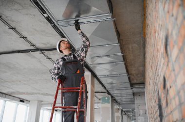 Güvenlik miğferi olan bir inşaatçı inşaat halindeki bir binada merdiven kullanarak havalandırma sistemi kuruyor.