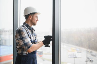 Güvenlik kaskı takan ve inşaat halindeki bir binaya pencere çerçeveleri yerleştirmek için elektrikli tornavida kullanan inşaatçı.