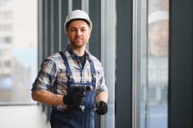Güvenlik kaskı takan inşaat işçisi ve modern bir binaya cam yerleştirirken kablosuz matkap tutuyor.