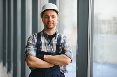 Modern binada kolları çaprazlanmış vaziyette duran kasklı ve iş üniformalı gülümseyen hvac teknisyeninin portresi.