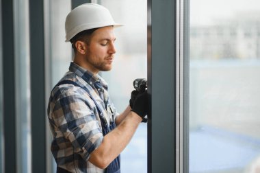 Koruyucu kask ve eldiven giyen inşaatçı kablosuz matkap kullanarak modern binaya pencere çerçevesi kuruyor.