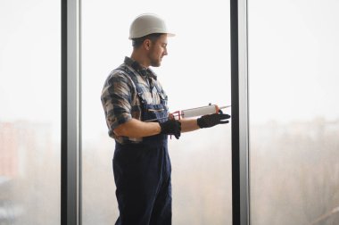 İnşaat işçisi, güvenlik kaskı ve eldiven takarak inşaat halindeki bir binanın pencere çerçevesine dolgu macunu sürüyor.