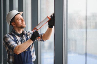 İnşaat işçisi, güvenlik kaskı ve eldiven takarak inşaat halindeki bir binanın pencere çerçevesine dolgu macunu sürüyor.