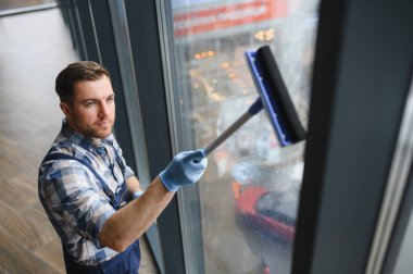 Profesyonel pencere temizleyicisi mavi eldiven takıyor. Modern bir binanın camını silmek için cam sileceği kullanıyor.