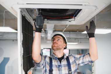 Güvenlik kaskı takan ve işçi tulumu giyen bir elektrikçi elektrikli matkap kullanarak havalandırma sistemi kuruyor.