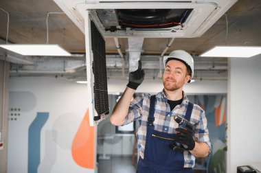 Elektrik teknisyeni, kablosuz tornavidayı tutarken tavana monte edilmiş klima ünitesini işaret ederek modern ofis alanında bakım veya onarım yapıyor.