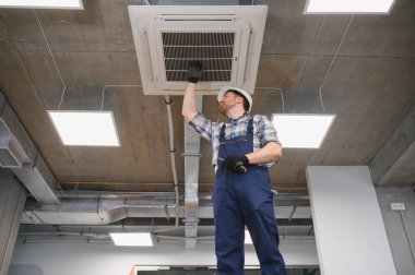 Kasklı ve işçi tulumlu elektrikçi bakım sırasında havalandırma ızgarasını kontrol ediyor.