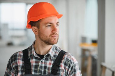 İnşaat işçisi turuncu bir şapka takıyor ve inşaat halindeki bir binayı inceliyor.