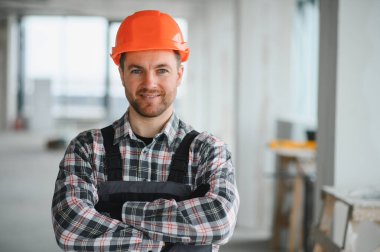 Turuncu kasklı ve tulumlu gülümseyen bir inşaat işçisinin portresi inşaat halindeki bir binada kolları çapraz duruyor.