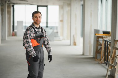 İnşaat işçisi inşaat halindeki binada miğfer ve iş tulumu ve eldiven giyerek yürüyor.