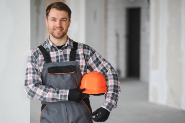 İnşaat halindeki bir binada kolunun altında turuncu bir miğfer tutan gülümseyen bir inşaat işçisinin portresi.