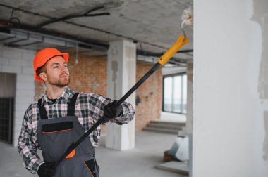 İnşaat işçisi koruyucu kask ve tulum giyiyor, uzun bir silindir kullanarak duvarı boyuyor, tadilat sırasında...