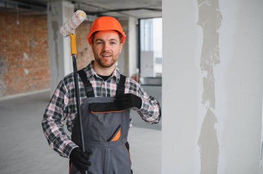 Tulumlu, kasklı, elinde boya silindiri olan ve kendini gösteren kendine güvenen inşaat işçisi inşaat halindeki bir binada tadilata başlamaya hazır.