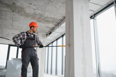 İnşaat işçisi koruyucu kask ve tulum giyiyor, uzun bir silindir kullanarak duvarı boyuyor, tadilat sırasında...