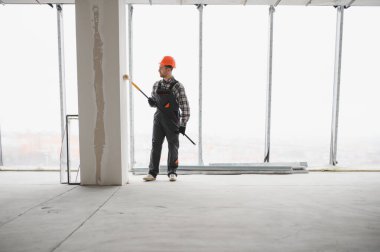 İnşaat işçisi, güvenlik kaskı takarken silindirle duvar boyuyor ve modern bir inşaat alanının yenilenmesine katkıda bulunuyor.