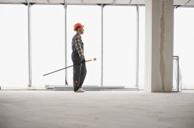 İnşaat işçisi inşaat halindeki bir binada yürüyor, uzun bir boya silindiri taşıyor ve boyama işine hazırlanıyor.