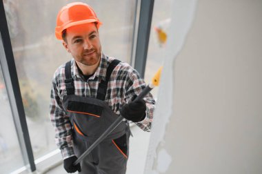 İnşaat işçisi koruyucu kask ve tulum giyiyor, uzun bir silindir kullanarak duvarı boyuyor, tadilat sırasında...