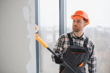 Koruyucu kask ve tulum giyen inşaat işçisi teleskopik boya silindiri kullanıyor, büyük bir pencerenin yakınındaki duvarı boyuyor.