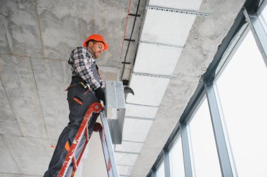 İnşaat işçisi miğfer takıyor. İnşaat halindeki bir binanın merdivenine havalandırma boruları yerleştiriyor.