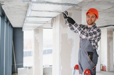 Güvenlik kaskı ve tulum giyen inşaat işçisi, inşaat halindeki binanın tavanına havalandırma sistemi kurmak için aletler kullanıyor.