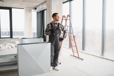 İnşaat işçisi tadilat altındaki bir binaya havalandırma kuruyor, elinde kablosuz bir tornavida tutuyor ve yan bakıyor.