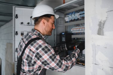 Elektrik paneli üzerinde çalışan, güvenlik kaskı takan ve alet kullanan, bakım ve onarım yapan bir elektrikçi.