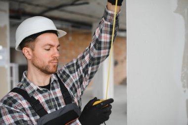 İnşaat işçisi güvenlik kaskı ve eldiven takıyor ve bina tadilatı sırasında mezura kullanıyor.