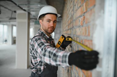 İnşaat alanında tuğla duvarda çalışırken kask ve eldiven takan kişi mezura ve ruh seviyesini kullanıyor.