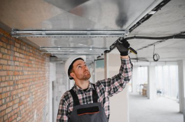 Güvenlik kaskı takan ve tulum giyen inşaat işçisi inşaat halindeki binanın tavanına havalandırma boruları yerleştiriyor.