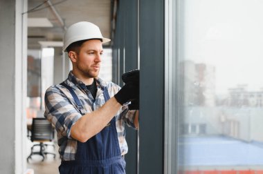 Güvenlik kaskı takan ve iş tulumu giyen profesyonel müteahhit, inşaat halindeki modern bir binaya pencere takmak için kablosuz tornavida kullanıyor.