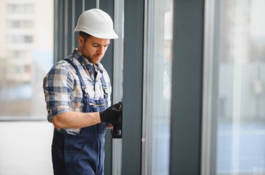 Koruyucu kask ve eldiven takan konsantre inşaat işçisi, yenilenme sırasında modern binaya pencere çerçeveleri kuruyor