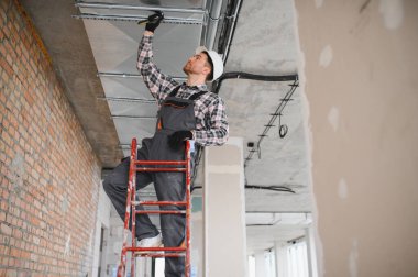 Güvenlik kaskı takan inşaat işçisi merdivenin üzerinde duruyor, inşaat halindeki bir binanın havalandırma sistemini tamir ediyor.