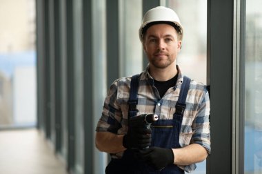 İnşaat işçisi güvenlik kaskı takıyor ve inşaat halindeki pencerenin yanında dururken kablosuz matkap tutuyor.