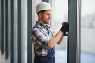 Güvenlik kaskı ve eldiven takan inşaat işçisi, modern bir binaya pencere çerçevesi takmak için elektrikli tornavida kullanıyor.