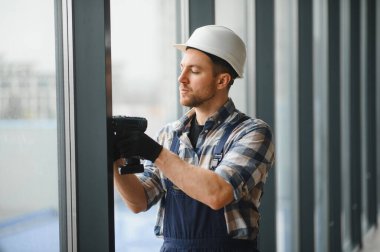 Güvenlik kaskı takan ve kablosuz tornavida kullanan inşaat işçisi, inşaat halindeki modern bir binaya pencere takıyor.