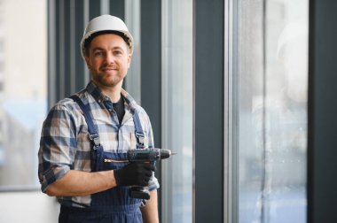 İnşaat işçisi güvenlik kaskı takıyor ve bir binaya cam yerleştirirken kablosuz matkap tutuyor.