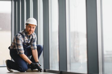 Kablosuz matkap tutan inşaat işçisi, modern bir ofis binasında büyük bir pencerenin önünde diz çökmüş, onarıma veya kuruluma hazır.