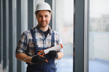 Koruyucu kask ve eldiven takan inşaat işçisi, silikon dolgu tabancası tutuyor, modern binaya pencere kurmaya hazırlanıyor.