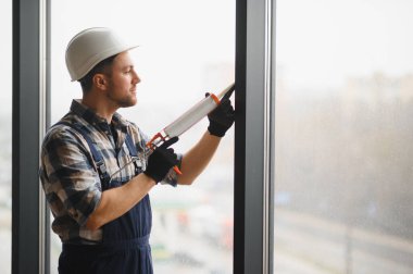 İnşaat işçisi koruyucu kask ve eldiven takıyor ve bina tadilatı sırasında pencere çerçevesine dolgu macunu sürüyor.
