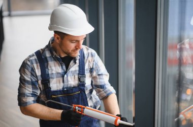İnşaat işçisi, güvenlik kaskı ve eldiven takarak inşaat halindeki bir binanın pencere çerçevesine dolgu macunu sürüyor.