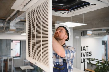 Güvenlik kaskı takan HVC teknisyeni ofis binasında tavan monte edilmiş havalandırma ünitesini tamir ediyor.