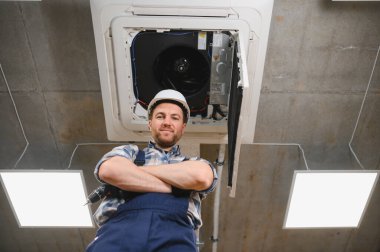 Kollarını kavuşturmuş, iş tulumu ve kasketi takmış, yeni kurulmuş havalandırma ünitesinin altında duran kendinden emin bir HVK teknisyeni.