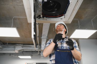 Elektrikçi, bir tornavidayla özenle çalışırken güvenlik için koruyucu bir başlık ve eldiven takarken bir havalandırma sistemini tamir ediyor.