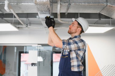 İnşaatçı güvenlik kaskı takıyor ve ticari bir alana ustalıkla bir havalandırma sistemi kurarken çeşitli aletler kullanıyor.